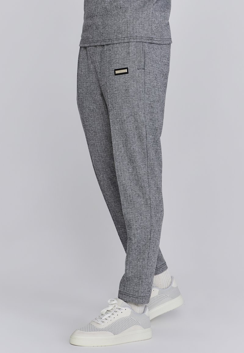 Pantalon gris à motif chevrons porté avec des chaussettes blanches et des baskets blanches sur une personne debout devant un fond uni.