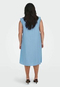 Světle modré šaty bez rukávů s volánkovými detaily na ramenou, hladký materiál, délka ke kolenům a uvolněný střih, v kombinaci s černými sandály na podpatku s otevřenou špičkou.