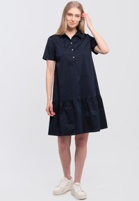 Navyblauw korter mouwjurk met een kraag, knopenrij en gerimpelde zoom. Gemaakt van een soepele stof, gecombineerd met witte sneakers.