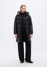 Μαύρο μπουφάν γεμισμένο με κουκούλα, με σχεδιασμό καπιτονέ, λαμπερή υφή και μακρύ μήκος. Φορεμένο πάνω από μαύρο πλεκτό πουλόβερ και παντελόνια.