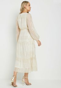 Largo vestido color crema con mangas transparentes y texturizadas y rayas doradas verticales. Presenta una falda con volantes y una cintura ceñida. Se lleva con sandalias de tacón doradas.