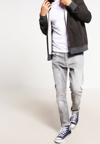 Felicissimo hoodie grigio con zip sopra una maglietta bianca, jeans grigio chiaro e sneaker blu scuro. L'outfit presenta un design casual e una vestibilità rilassata.
