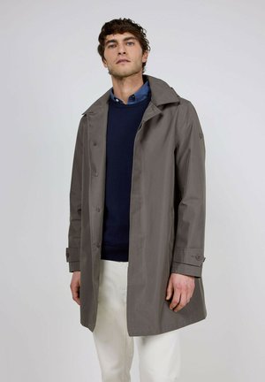 Hombre que lleva un abrigo tipo trench gris abotonado sobre un suéter azul marino y una camisa azul con cuello, de pie frente a un fondo claro y sencillo.