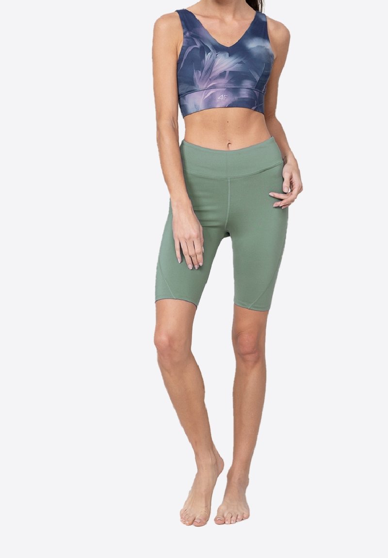 Kvinde iført en lilla mønstret sport-bh og højttaljede grønne cykeltights, stående barefodet mod en ensfarvet baggrund.
