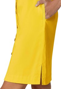 Gelbes Baumwollkleid mit knielangem Schnitt, vorderen Knöpfen, seitlichen Taschen und einem kleinen Seitenschlitz für Bewegungsfreiheit.