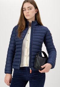 Marineblauwe pufferjack met horizontale stiknaad, hoge kraag, oranje ritshendel, gedragen over een crèmekleurige kabelgebreide trui en gecombineerd met donkerblauwe corduroy broek.