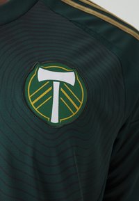 Tummanvihreä urheilupaita, jossa on kapeita mustia vaakaraitoja. Paidassa on pyöreä emblem valkoisesta kirveestä sekä vihreistä ja keltaisista geometrisista kuvioista.