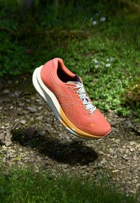 Chaussure de course orange avec une tige en mesh texturé, semelle intermédiaire blanche, lacets bleu clair et accents jaunes subtils, positionnée au-dessus d'un sol rocheux.