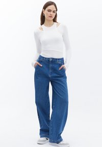 Top blanco de manga larga con detalle de hombro descubierto, combinado con jeans anchos de tiro alto azul. Modelo con las manos en los bolsillos.