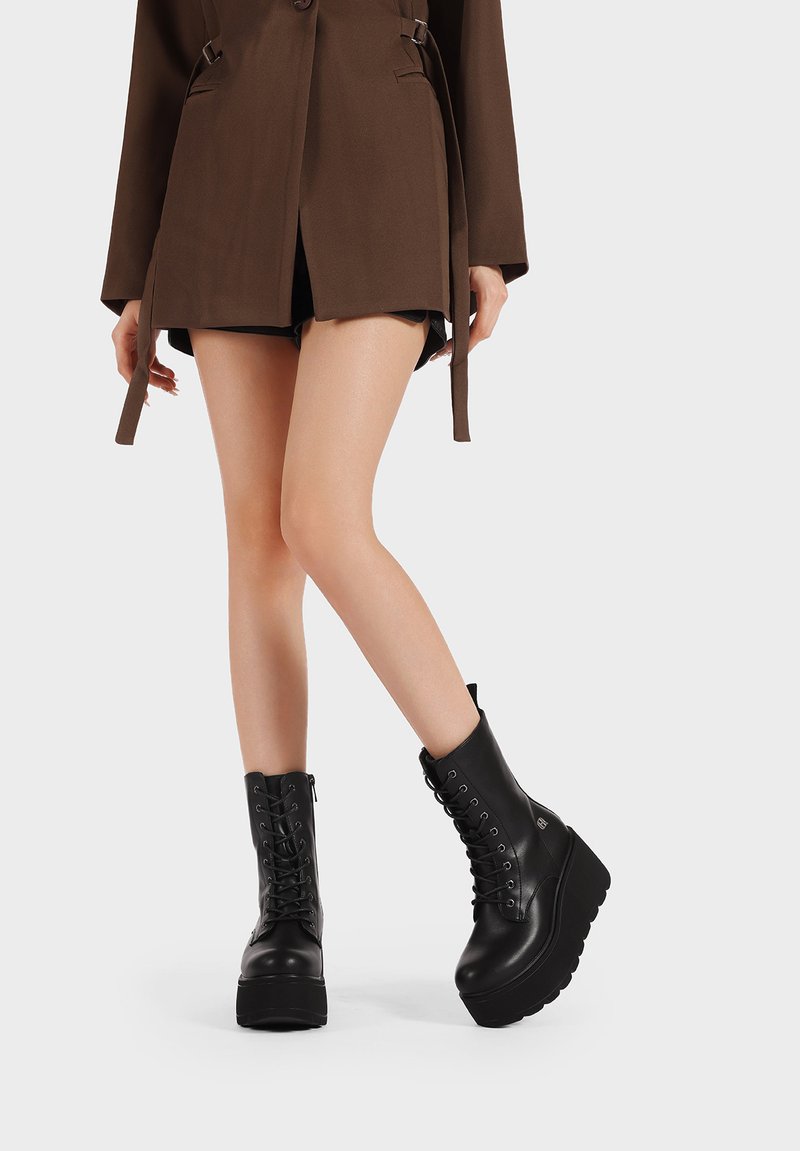 Botas de combate negras con plataforma, cordones en la parte delantera, suela gruesa, textura de cuero suave y detalles de costura. Llevadas con una chaqueta marrón.