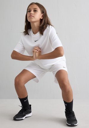 Hvit idrettsdrakt og shorts med svart Nike-logo, svarte sokker og svarte joggesko; person som croucher i en treningsposisjon.