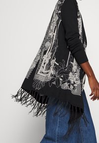 Svart sjal med vita blommönster och geometriska mönster, med fransiga kanter och lös passform. Tillverkad av mjukt, strukturerat tyg.