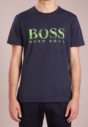 Mann trägt ein marineblaues Kurzarm-T-Shirt mit großem grünem "BOSS HUGO BOSS" Logo auf der Brust.