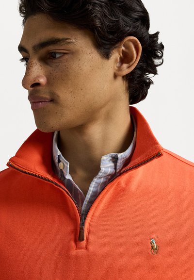 Pulover fermoar portocalie cu material texturat, având un logo pe pieptul stâng și o cămașă în carouri albastre și albe dedesubt.