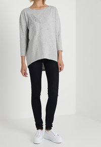 Sudadera grande de color gris claro con escote barco y un detalle en el lado. Combinada con jeans ajustados oscuros y zapatillas blancas con plataforma.