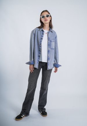 Světlá modrá džínová overshirt s kapsou, vrstvená přes bílý tričko, spárována s tmavě šedými kalhotami a černými botami s bílými pruhy.