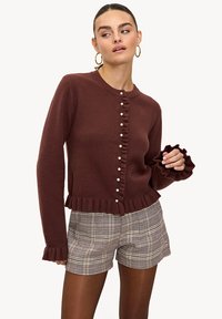 Cardigan en tricot marron avec un col rond, un ourlet à volants et une fermeture à boutons, associé à un short à carreaux. Tissu texturé.