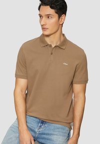 Camisa polo marrón con un acabado texturizado, que presenta un cuello con dos botones y un pequeño logo blanco en el lado izquierdo del pecho. Mangas cortas, ajuste casual.