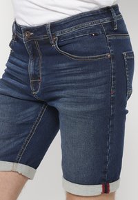 Jeans šorts u tamnoplavom pranju s svijetloplavim preklopom. Ima stilsku šavovu obradu i mali crveni detalj blizu džepa.