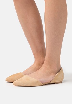Chaussures plates en daim beige clair avec un bout pointu et des côtés ouverts, présentant une texture lisse et un design minimaliste.