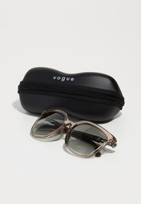 Ochelari de soare Vogue cu rame supradimensionate, transparente, de culoare maro și lentile gri, așezati lângă o husă neagră cu fermoar care poartă numele brandului.