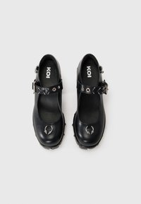 Black patent leather Mary Jane shoes with a strap, silver buckle, and decorative emblem on the toe. Features a textured insole and studded accents.