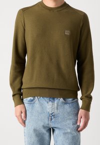 Sweter oliwkowozielony o teksturowanym splocie, z prążkowanym kołnierzem i mankietami, z małą naszywką z logo na piersi. Zestawiony z jasnoniebieskimi jeansami.