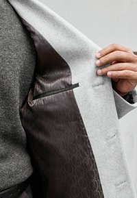 Abrigo gris claro con un interior suave que presenta un forro de patrón oscuro y un detalle sutil en los bolsillos, resaltando el contraste de telas y el diseño.