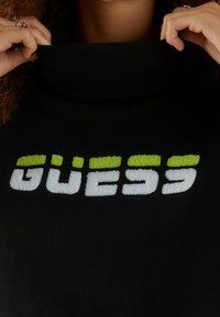 Pull noir avec un col montant. Le devant affiche le logo "GUESS" texturé en blanc et vert vif. Tissu lisse dans l'ensemble.