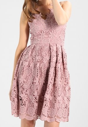 Kvinde iført en ærmeløs støvet lyserød blondekjole med blomsterprint og fitted talje, stående mod en ensfarvet lys baggrund.