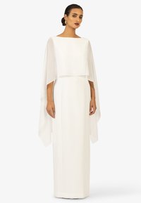 White gown featuring a boat neckline and layered sheer cape sleeves. The silhouette is sleek and floor-length with a smooth texture.