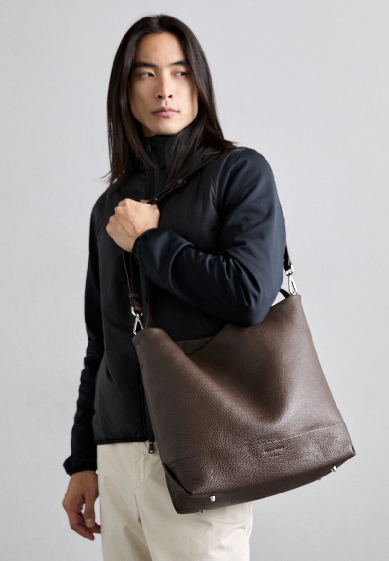 Bolsa de ombro em couro castanho com um design relaxado, superfície texturizada, com detalhes em metal prateado e tachas laterais, transportada com uma alça destacável.