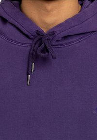 Primer plano de una persona que lleva una sudadera morada con un cordón atado en el cuello y una textura de tela suave.