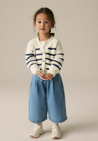 Strickjacke mit cremefarbener Basis, dunkelblauen Streifen und floralen Akzenten. Breite Denim-Culottes, cremefarbene Socken und beigefarbene Sneaker runden das Outfit ab.