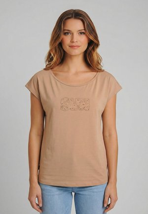 Donna con capelli di media lunghezza che indossa una maglietta beige con la scritta "SUN" e jeans azzurri, in piedi su uno sfondo grigio semplice.