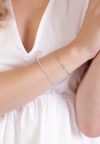Des bracelets en argent avec des maillons circulaires et ovales, l'un orné de pierres claires, portés sur un bras à la peau claire sur fond blanc.