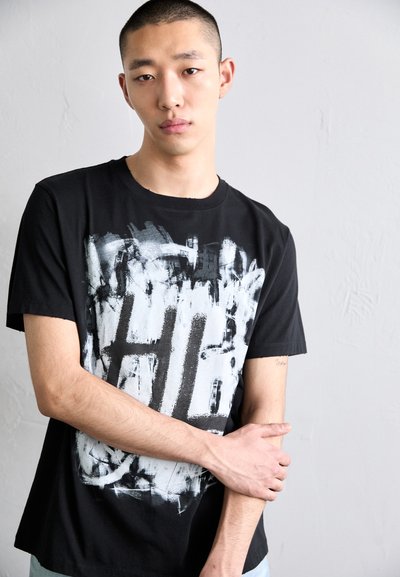 Fekete rövid ujjú póló, amelyen fehér és szürke absztrakt minta található a mellkas területén. Pamut anyag, kerek nyakkivágás, lazább szabás.