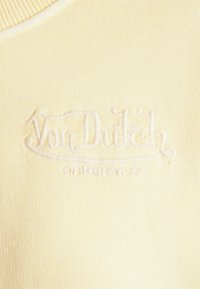 Žlutá pletenina se vyšívaným logem "Von Dutch" v bílé barvě, umístěným na středu. Hladká textura s žebrovaným výstřihem.