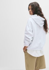 Felpa grigia con cappuccio, vestibilità ampia, polsini a coste. Indossata sopra una camicia bianca a righe. Abbinata a pantaloni corti beige, mostrando un outfit casual.