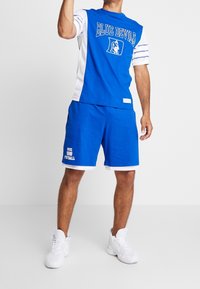 Hombre vistiendo una camiseta deportiva "Blue Devils" en azul y blanco y pantalones cortos a juego, con zapatillas deportivas blancas, levantando un brazo.