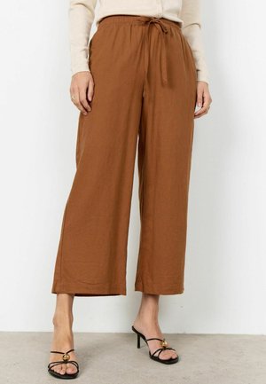 Donna che indossa pantaloni marroni a gamba larga e taglio corto con vita regolabile e sandali neri con tacco e dettagli dorati, in piedi su pavimento di cemento.