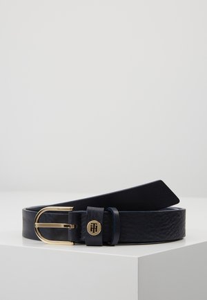 Ceinture en cuir noir avec une boucle ronde dorée et un petit emblème rond en or portant les initiales TH sur la passants de ceinture.
