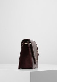 Vinrød læderhåndtaske med en struktureret form, der har guldfarvede nitter langs kanten og en klaplukning. Blød tekstur med minimalt hardware.