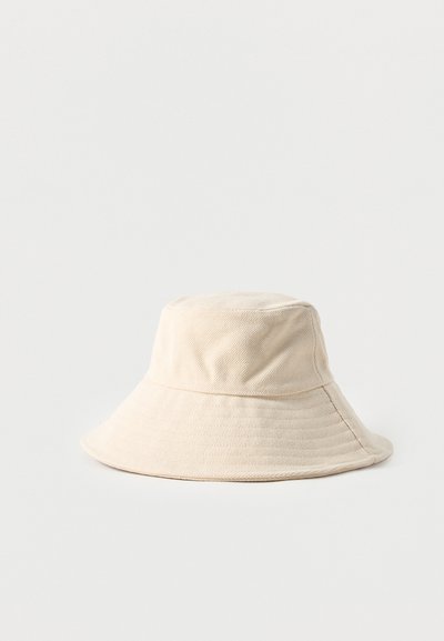 Μπεζ βαμβακερό καπέλο τύπου bucket με φαρδύ, μαλακό γείσο και ορατές ραφές σε απλό, ανοιχτό χρώμα φόντο.