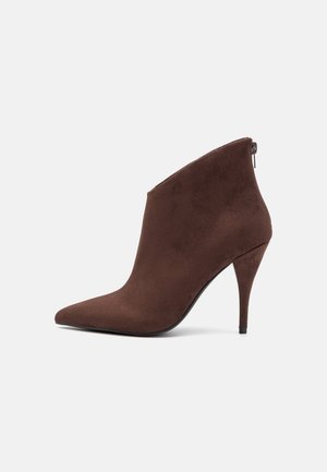Brun ruskindsankelstøvle med spids snude, stiletthæl og lynlås bagpå på lys baggrund.