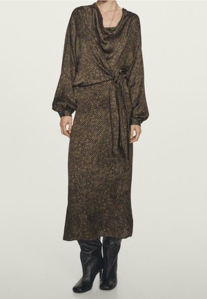 Femme portant une robe midi marron à manches longues avec un motif, un lien sur le côté et des bottes noires à talons sur un fond uni.
