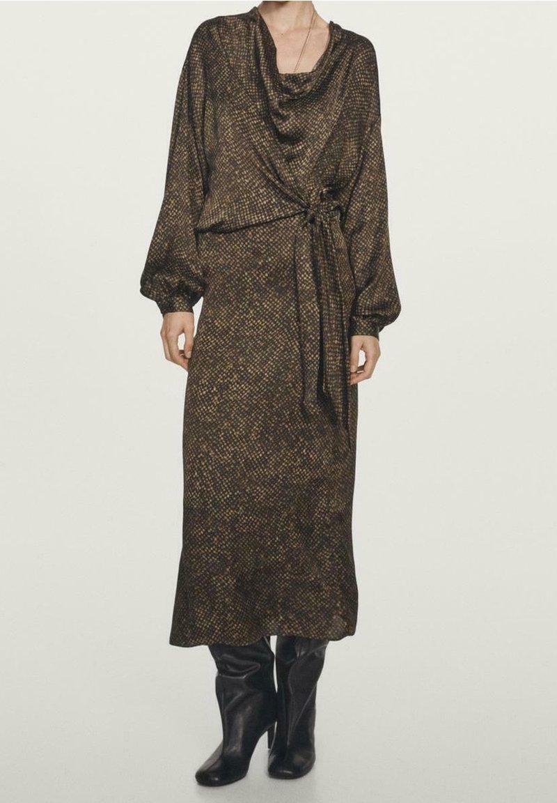 Femme portant une robe midi marron à manches longues avec un motif, un lien sur le côté et des bottes noires à talons sur un fond uni.