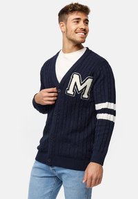 Marineblå kabelstrikket cardigan med V-udskæring, prydet med en hvid bogstav 'M' patch og stribede hvide detaljer på ærmerne, afsluttet med knaplukning.