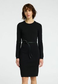 Robe en maille noire à manches longues, décolleté rond, silhouette ajustée, et une ceinture en cuir verte nouée à la taille.