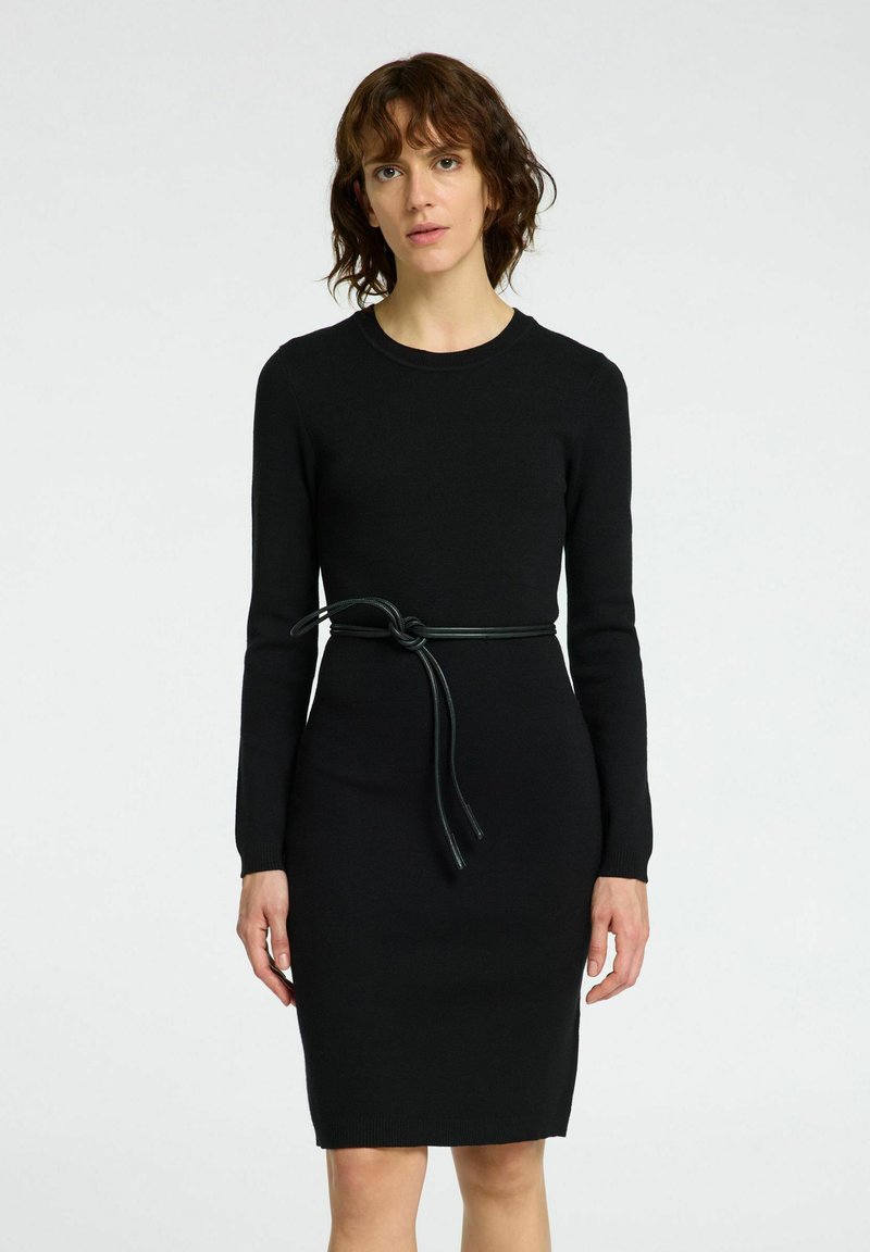 Zwarte gebreide jurk met lange mouwen, ronde halslijn, getailleerde silhouet en een groene leren riem die geknoopt is in de taille.